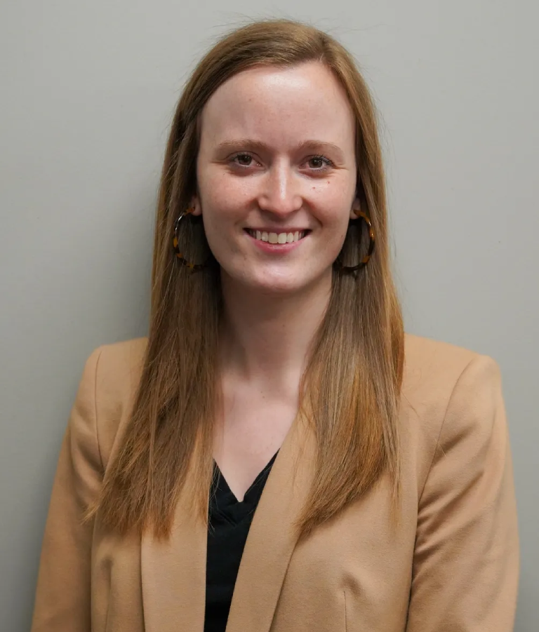 Smiling woman wearing a tan blazer and black top against a gray background.